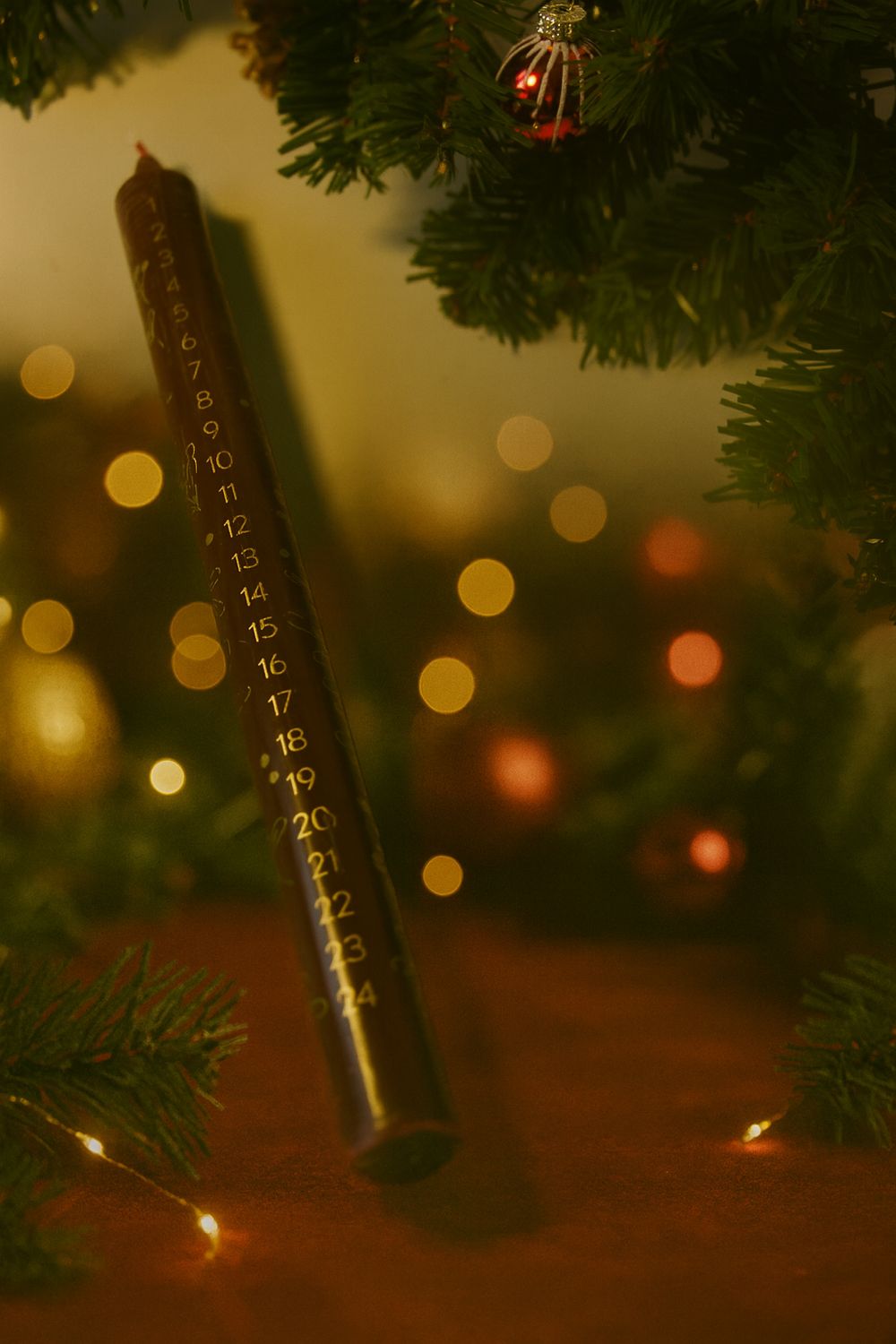 Holz-Adventskalender mit Zahlen neben grünem Kiefernzweig, roten Weihnachtskugeln und warmweißer Lichterkette im Hintergrund.