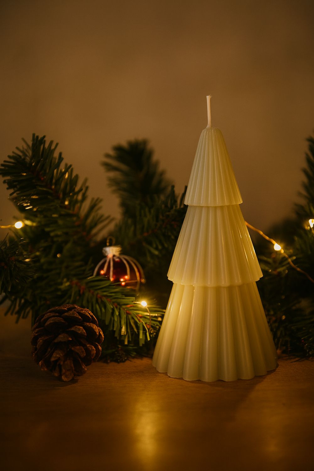 Cremefarbene Weihnachtskerze in Tannenbaum-Form neben Pinecone und Evergreens mit warmem Licht.